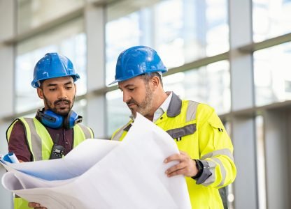 Young indian architect and mature supervisor meeting at construction site. Multiethnic manual worker and engineer wearing safety equipment and discussing on plan. Two construction workers working together while visiting a new building.